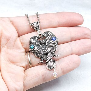 May include: Silver necklace with a pendant of two birds facing each other, their wings forming a heart shape. The birds have blue and green gemstone eyes and a crystal dangle.