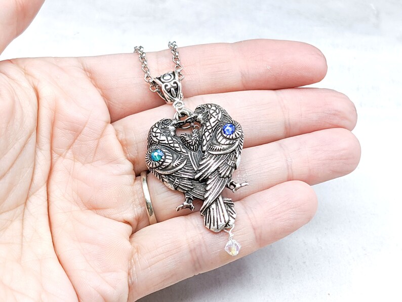 May include: Silver necklace with a pendant of two birds facing each other, their wings forming a heart shape. The birds have blue and green gemstone eyes and a crystal dangle.