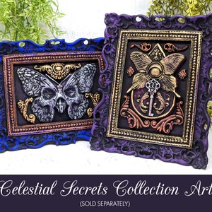 May include: Two framed pieces of art with a celestial theme. The first frame is blue and features a silver butterfly with a skull head. The second frame is purple and features a gold butterfly with a key in the center. Both frames have ornate gold trim.