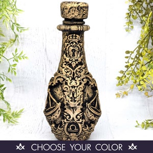 May include: A decorative glass bottle with an ornate design and a gold finish. The bottle features the astrological symbol for Libra and a keyhole design.