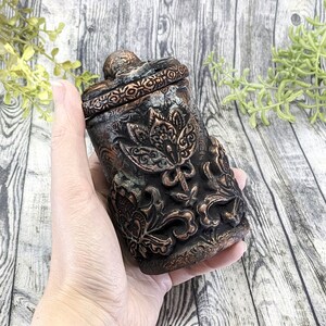 May include: A small, ornate, black and copper-colored ceramic jar with a floral design. The jar has a lid and is held in a hand.