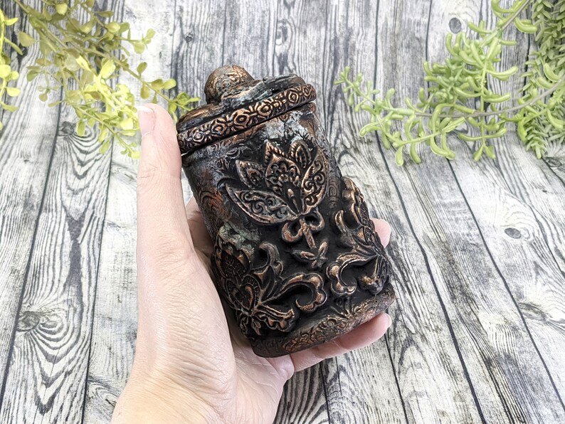 May include: A small, ornate, black and copper-colored ceramic jar with a floral design. The jar has a lid and is held in a hand.