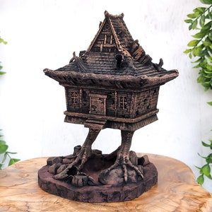 May include: A small, wooden house with a shingled roof sits on two bird-like legs. The house is painted in a dark brown color and has a small door and windows. The house is sitting on a brown, circular base.