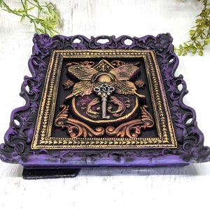 May include: A purple and gold ornate frame with a decorative design featuring a key and butterfly wings. The frame is made of resin and has a distressed finish.