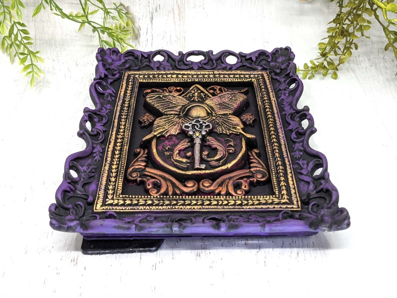 May include: A purple and gold ornate frame with a decorative design featuring a key and butterfly wings. The frame is made of resin and has a distressed finish.