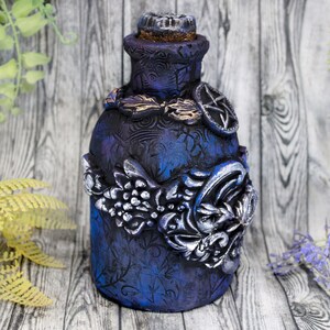 May include: A dark blue glass bottle with silver and gold embellishments. The bottle has a textured surface and is decorated with floral and geometric designs.