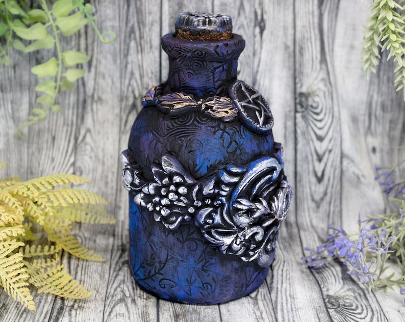 May include: A dark blue glass bottle with silver and gold embellishments. The bottle has a textured surface and is decorated with floral and geometric designs.