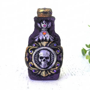 May include: A small purple glass bottle with a gold and silver skull design. The bottle has a gold top and is decorated with purple gems.