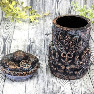 May include: A decorative ceramic jar with a lid. The jar is brown and copper colored with intricate floral designs. The lid has a similar design.