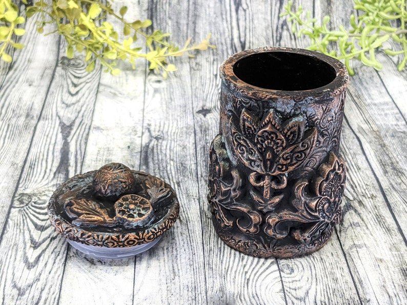 May include: A decorative ceramic jar with a lid. The jar is brown and copper colored with intricate floral designs. The lid has a similar design.