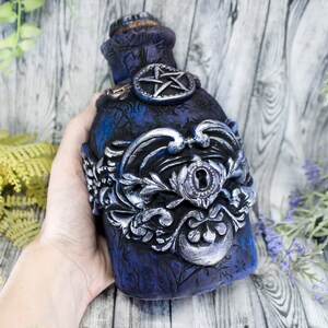 May include: A dark blue glass bottle with a silver metal design and a keyhole. The bottle has a pentagram design on the front.