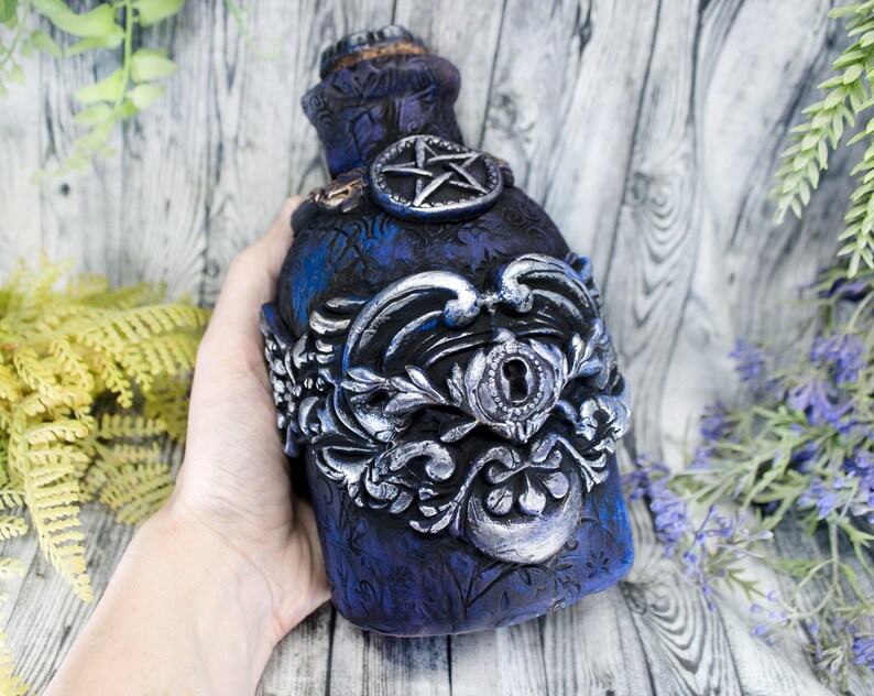May include: A dark blue glass bottle with a silver metal design and a keyhole. The bottle has a pentagram design on the front.
