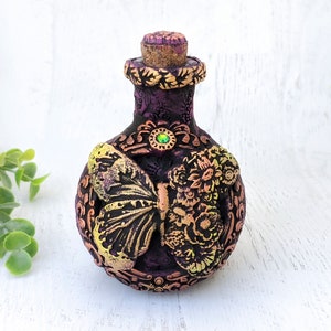 以下が含まれることがあります： コルク栓と金の蝶のデザインが施された、小さな紫色のガラス瓶。瓶は複雑な花の模様と緑色の宝石で飾られています。