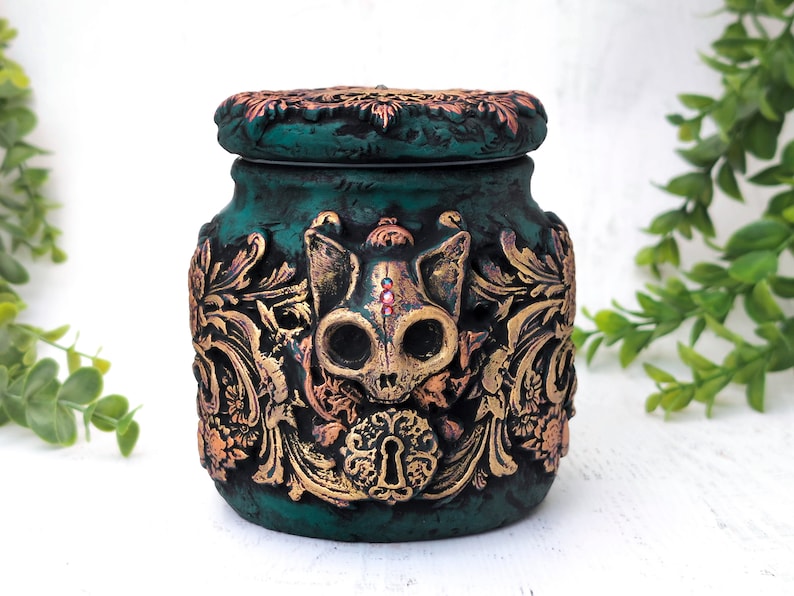 May include: A teal green ceramic jar with a gold and copper lid. The jar is decorated with a skull of a cat with gold accents and a keyhole design. The jar has a distressed look.
