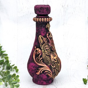 May include: A decorative ceramic bottle with a gold wolf design and intricate details. The bottle is painted in shades of purple and gold.