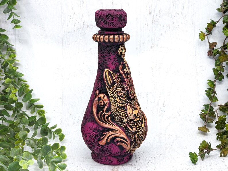 May include: A decorative ceramic bottle with a gold wolf design and intricate details. The bottle is painted in shades of purple and gold.
