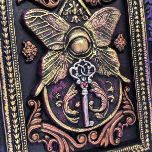 May include: A decorative framed artwork featuring a gold and purple butterfly with a silver key in the center. The frame is ornate and has a purple and gold design.