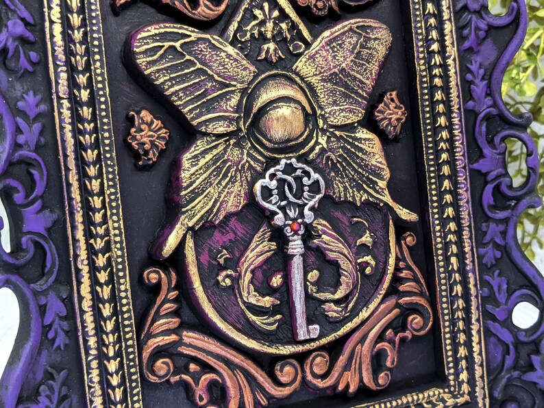 May include: A decorative framed artwork featuring a gold and purple butterfly with a silver key in the center. The frame is ornate and has a purple and gold design.