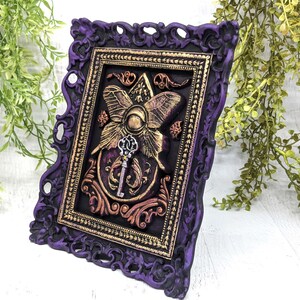 May include: A purple and gold framed wall art piece with a 3D butterfly and key design. The butterfly is gold with a purple center and the key is silver.