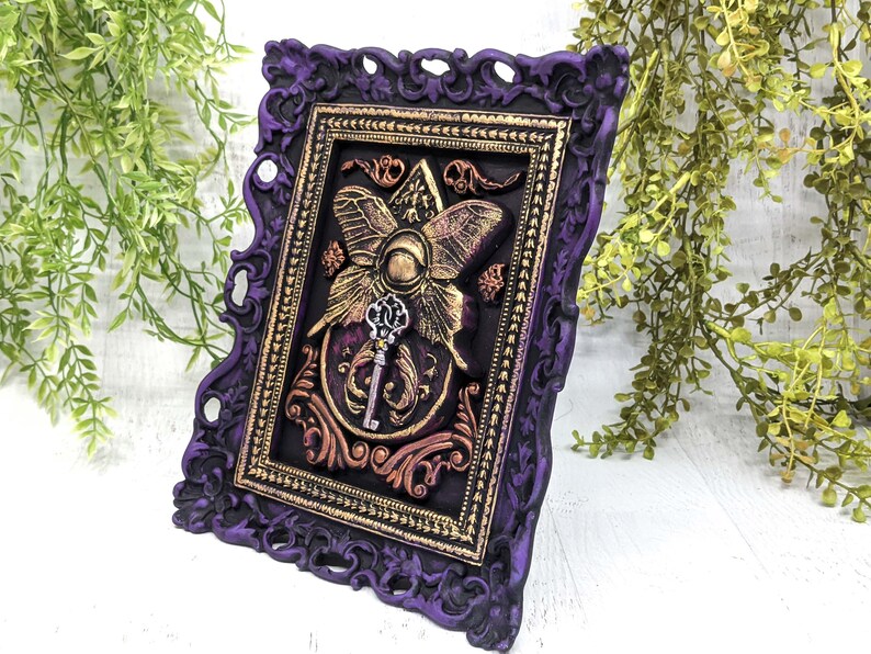 May include: A purple and gold framed wall art piece with a 3D butterfly and key design. The butterfly is gold with a purple center and the key is silver.