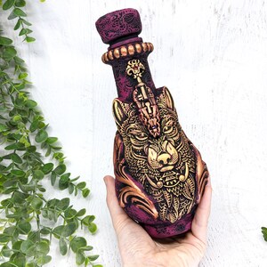 May include: A ceramic bottle with a wolf's head design. The bottle is painted in shades of purple and gold, with intricate details. The wolf's head is detailed with gold accents and a jeweled eye.