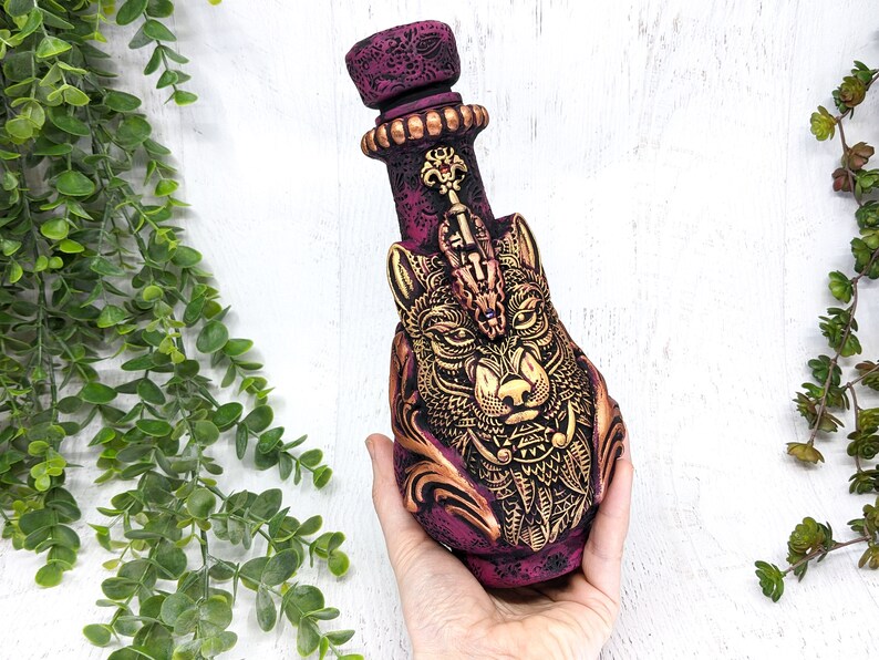 May include: A ceramic bottle with a wolf's head design. The bottle is painted in shades of purple and gold, with intricate details. The wolf's head is detailed with gold accents and a jeweled eye.