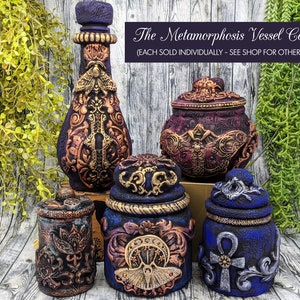 May include: A collection of five decorative vessels with intricate designs. The vessels are made of dark blue, purple, and gold paint with a variety of embellishments, including butterflies, keys, and an ankh symbol. The text "The Metamorphosis Vessel Collection (EACH SOLD INDIVIDUALLY - SEE SHOP FOR OTHER LISTINGS!)" is displayed above the vessels.