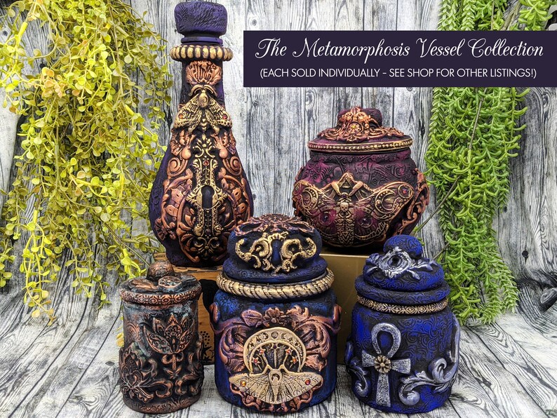 May include: A collection of five decorative vessels with intricate designs. The vessels are made of dark blue, purple, and gold paint with a variety of embellishments, including butterflies, keys, and an ankh symbol. The text "The Metamorphosis Vessel Collection (EACH SOLD INDIVIDUALLY - SEE SHOP FOR OTHER LISTINGS!)" is displayed above the vessels.