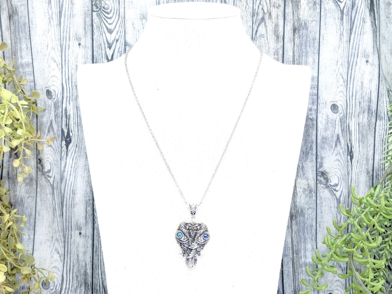 May include: A silver necklace with a detailed owl pendant featuring blue eyes. The owl pendant is a heart shape with intricate details.