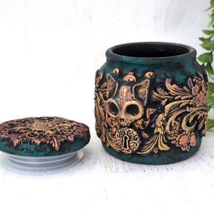 May include: Teal ceramic jar with a gold and copper skull design and a matching lid. The jar has a distressed finish and intricate details.