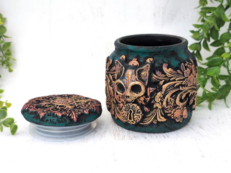 May include: Teal ceramic jar with a gold and copper skull design and a matching lid. The jar has a distressed finish and intricate details.
