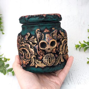 May include: Teal ceramic jar with a gold and copper skull design. The jar has a lid and a keyhole detail.