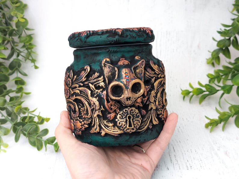 May include: Teal ceramic jar with a gold and copper skull design. The jar has a lid and a keyhole detail.