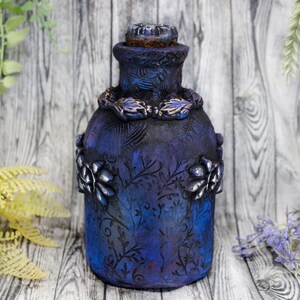 May include: A dark blue glass bottle with a decorative floral design and a cork stopper. The bottle has a textured surface and is decorated with silver accents.