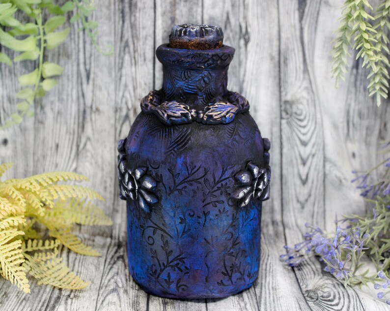 May include: A dark blue glass bottle with a decorative floral design and a cork stopper. The bottle has a textured surface and is decorated with silver accents.