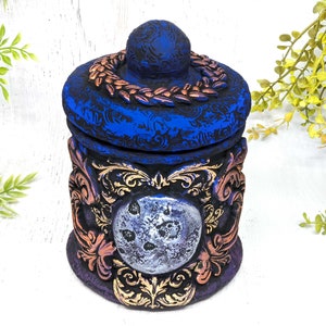 May include: A blue and gold ceramic jar with a moon design and ornate details. The lid has a wreath of leaves around the top.