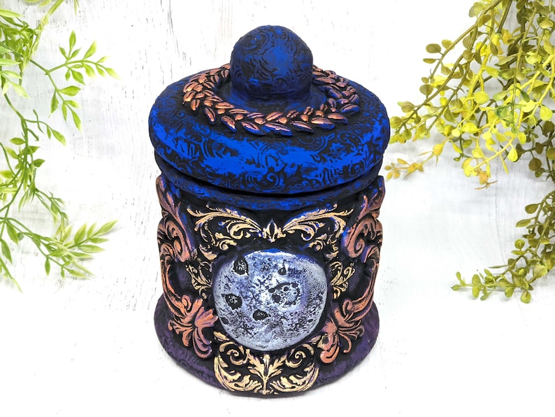 May include: A blue and gold ceramic jar with a moon design and ornate details. The lid has a wreath of leaves around the top.