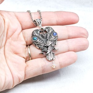 May include: Silver necklace with a pendant featuring two intertwined birds with blue gemstone eyes. The pendant has a crystal charm hanging from it.