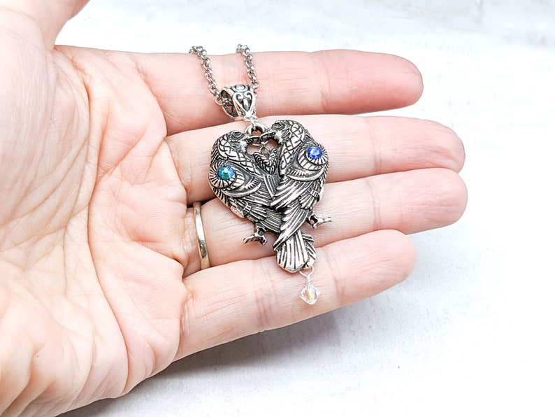 May include: Silver necklace with a pendant featuring two intertwined birds with blue gemstone eyes. The pendant has a crystal charm hanging from it.