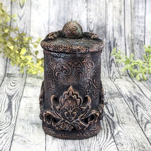May include: A decorative ceramic jar with an intricate floral design and a lid with a round knob. The jar is painted in a dark brown color with copper accents.