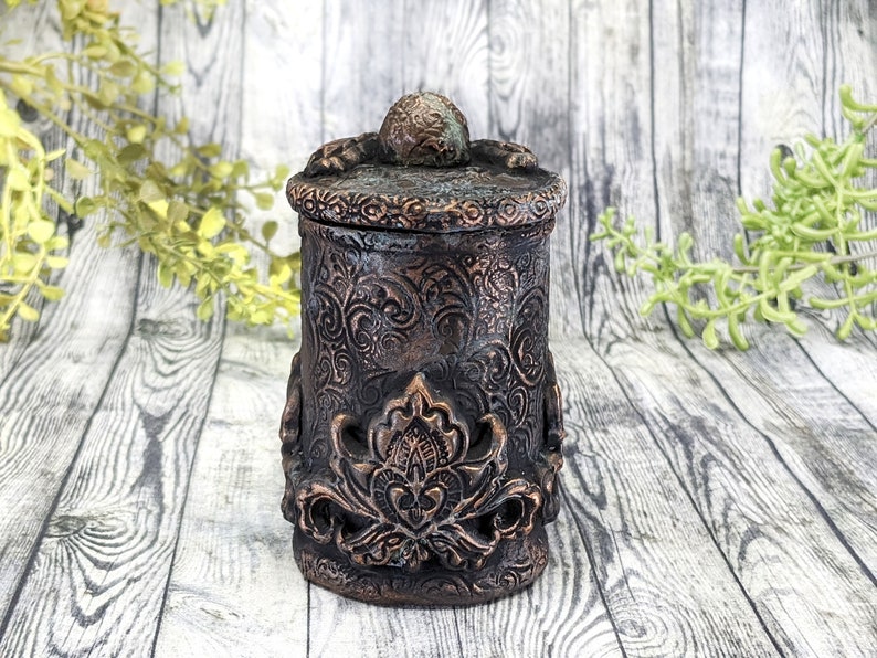 May include: A decorative ceramic jar with an intricate floral design and a lid with a round knob. The jar is painted in a dark brown color with copper accents.