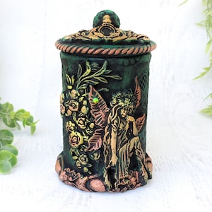 May include: A green ceramic jar with a lid, decorated with a fairy with wings, flowers, and gold accents.