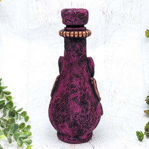 May include: A dark purple glass bottle with a decorative embossed design and a gold-colored stopper. The bottle is standing on a white surface.