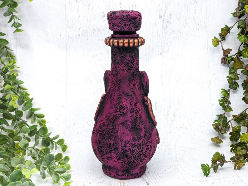 May include: A dark purple glass bottle with a decorative embossed design and a gold-colored stopper. The bottle is standing on a white surface.