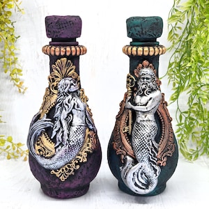 May include: Two decorative bottles with a mermaid and merman design. The bottles are painted in a purple and green color scheme with gold accents. The mermaid and merman are silver.