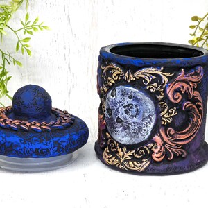 May include: A blue and purple ceramic jar with a gold and copper design. The jar has a lid with a copper band and a moon design. The jar is decorated with a moon and floral patterns.