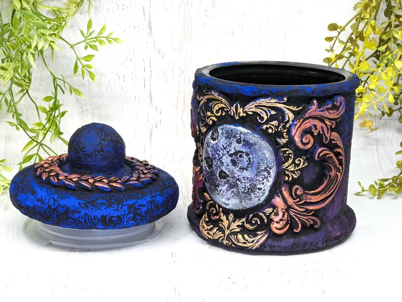 May include: A blue and purple ceramic jar with a gold and copper design. The jar has a lid with a copper band and a moon design. The jar is decorated with a moon and floral patterns.