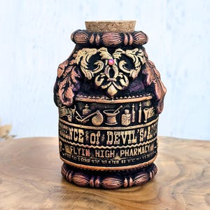 May include: An ornate, dark-coloured apothecary bottle with a cork stopper. The bottle features embossed details in gold and bronze, with the text "Essence of Devil's S***" and "Flyin' High Pharmacy". The design includes decorative leaves and apothecary symbols.