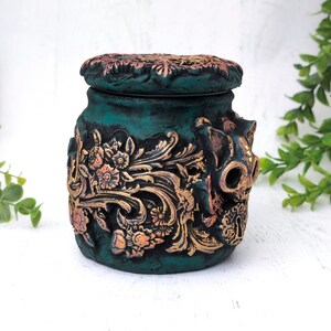 May include: A teal green ceramic jar with a gold floral design and a skull-shaped lid. The jar has a distressed finish and is decorated with gold accents.