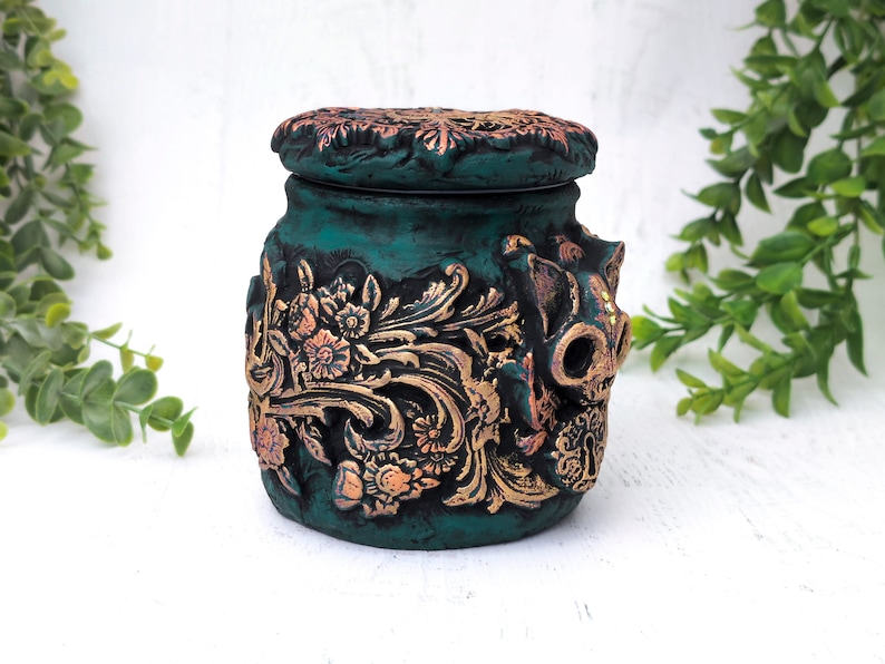 May include: A teal green ceramic jar with a gold floral design and a skull-shaped lid. The jar has a distressed finish and is decorated with gold accents.